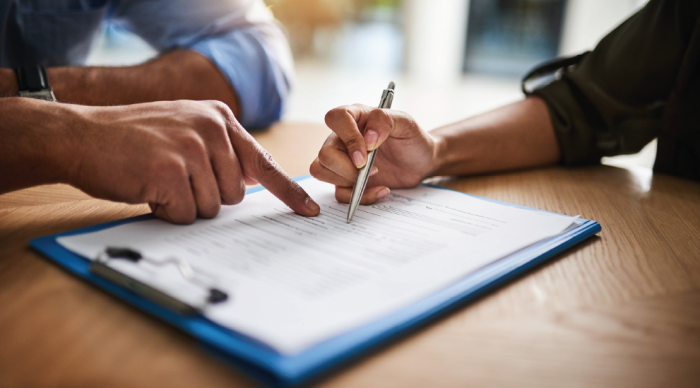  A man's hand points to a line on a document on a clipboard laying on a wooden table. A woman's hand holds a pen, poised to sign the document.  