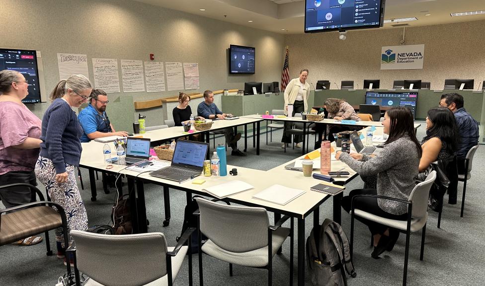 A group of adults sit and stand around U-shaped tables in a conference room, working on laptops and participating in a meeting. Papers, notebooks, water bottles, and small baskets are arranged on the tables. Posters with handwritten notes are taped to the wall, and large screens display a virtual meeting interface. A sign on the wall reads “Nevada Department of Education,” and an American flag stands near the front of the room.